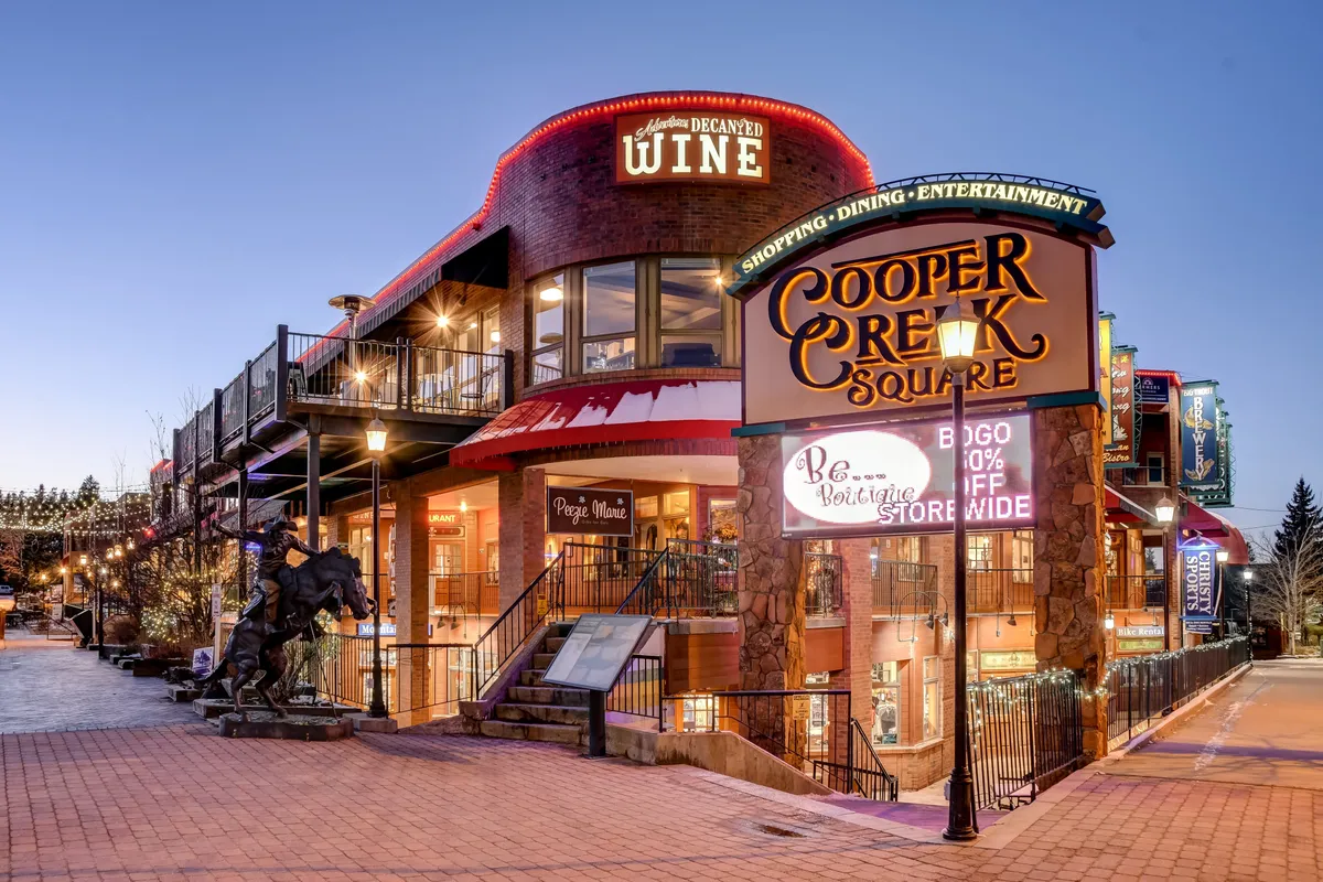 A view of Cooper Creek Square showcases its welcoming sign, wine shop, outdoor lighting, and sculpture. The scene is festive and inviting, capturing the ambiance of a lively local hub where shopping, dining, and entertainment converge.