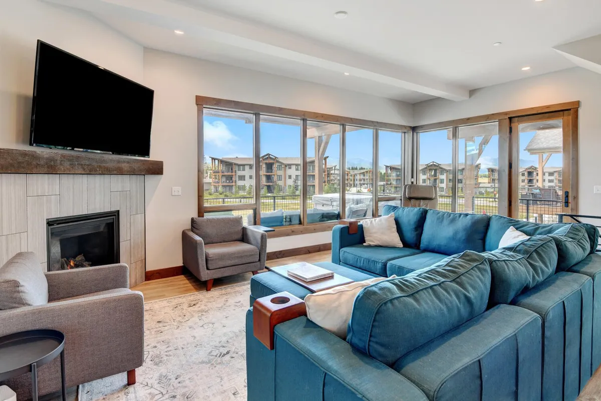 A spacious living room featuring a large sectional sofa arranged around a stone fireplace and a wall-mounted television. The room is filled with natural light from expansive windows that offer beautiful mountain views. The seating invites groups to gather for movies, conversation, or simply relaxing by the fire.
