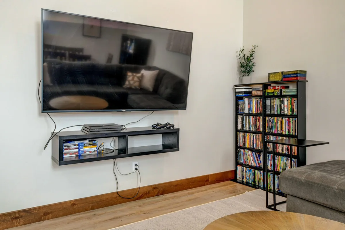 A cozy movie and game room featuring a wall-mounted TV, a shelf with a PlayStation 4 console and DVDs, and a plush, comfortable couch. The space is arranged for both viewing and playing, making it ideal for family game nights, movie marathons, or relaxing together after a day outdoors.