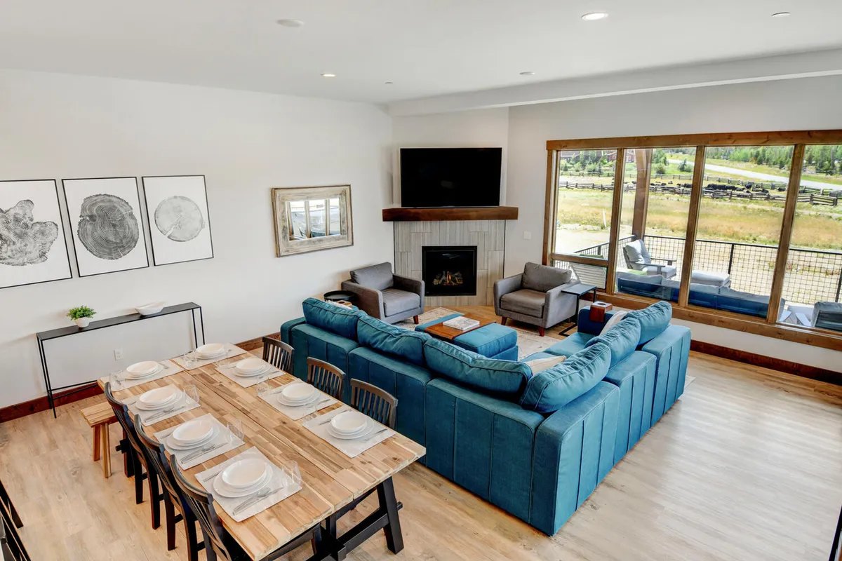 A cozy interior scene with a large sectional sofa, a dining table set for a meal, a fireplace, and large windows with a view. The space is designed for family gatherings, dinner parties, and relaxing movie nights, all with scenic views as a backdrop.