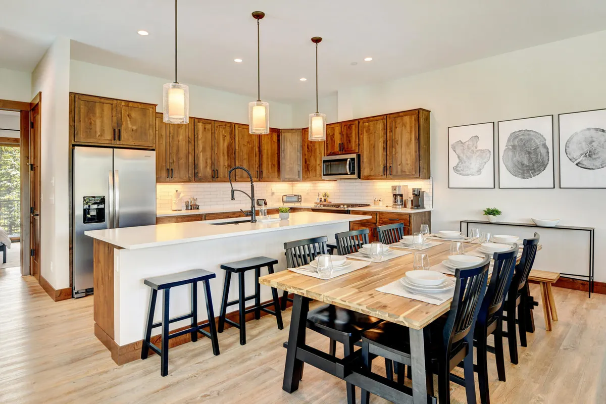 An open dining area with a large wooden dining table, a kitchen island in the background, and warm wood cabinetry. The space is set up for family-style meals or game nights, with artwork adding a personal, welcoming touch.
