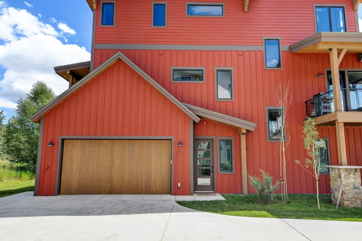 This image shows the vibrant red exterior of the house, featuring a wooden garage door, multiple windows, and a balcony area. The scene conveys an inviting and modern setting, with the balcony suggesting spaces for family gatherings and outdoor play.